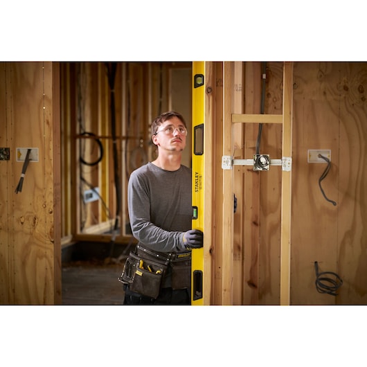 A person wearing work gloves and a tool belt is using a yellow Stanley spirit level to measure the vertical alignment of a wooden frame in a construction setting. The background shows unfinished wooden walls and electrical wiring.