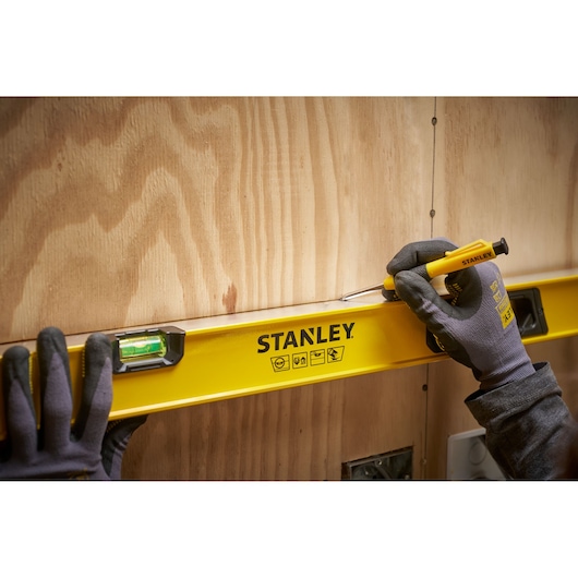 A person wearing gloves is using a yellow STANLEY spirit level on a wooden wall. The person is also holding a yellow screwdriver, marking or positioning along the edge of the level.