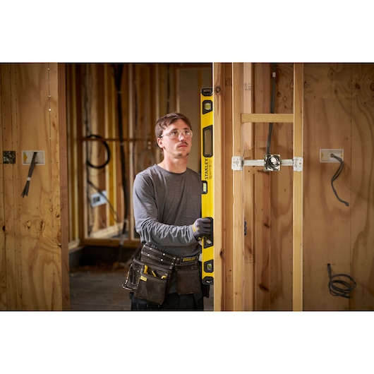 A person with their face blurred is holding a yellow STANLEY spirit level against a wooden wall frame on a construction site. The person is wearing work gloves and a tool belt with various tools.
