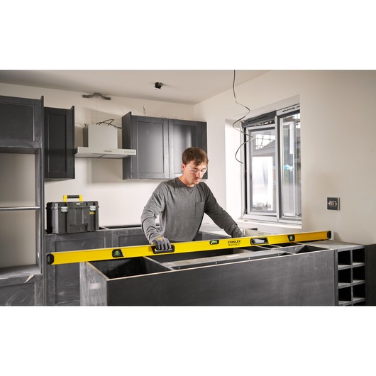 A person using a yellow STANLEY spirit level while working on a kitchen installation. The individual is wearing gloves and is measuring a countertop in a modern kitchen setting.