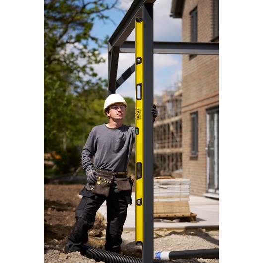 A construction worker in safety gear uses a yellow STANLEY spirit level (model STHT41778-0) to check the alignment of a vertical metal beam on an outdoor building site.