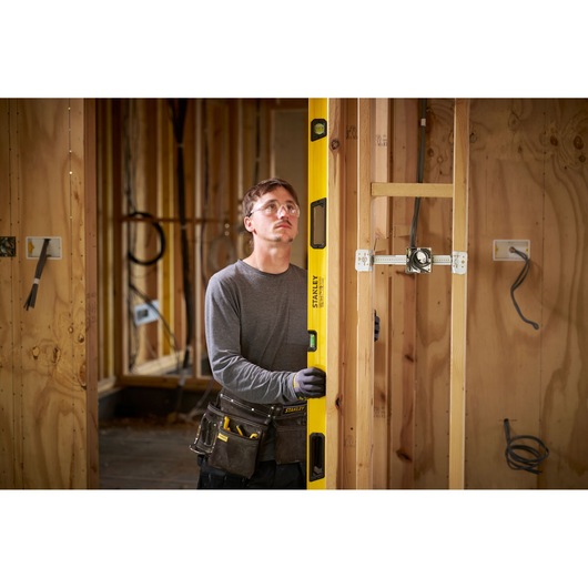 A person wearing a tool belt and gloves is using a yellow STANLEY spirit level to check the vertical alignment of a wooden frame in a construction site.