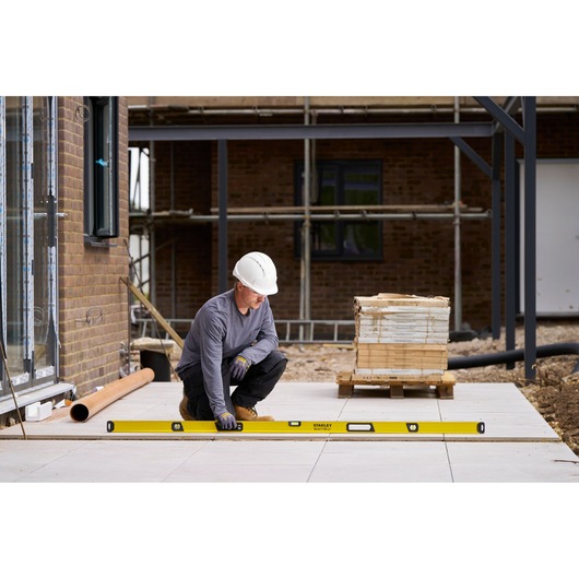 A construction worker wearing a white hard hat and gloves is kneeling outdoors, using a long yellow STANLEY spirit level to check the alignment of paving tiles. There are construction materials and scaffolding in the background.