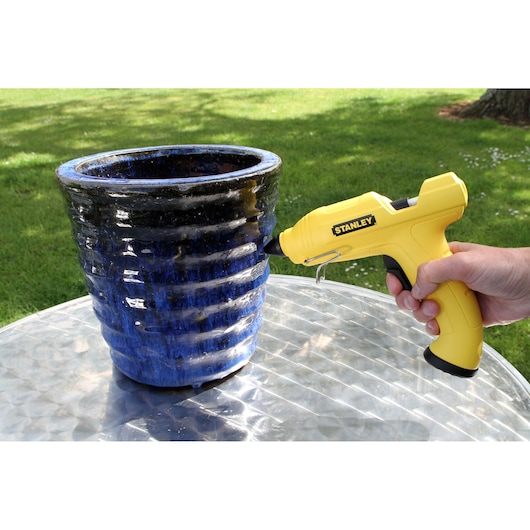 STANLEY STHT6-70416 yellow glue gun in use near a blue ceramic pot outdoors, right side view.