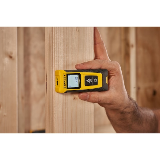 STANLEY STHT77065-0 laser distance measurer held against wooden beam, display screen lit, construction setting.