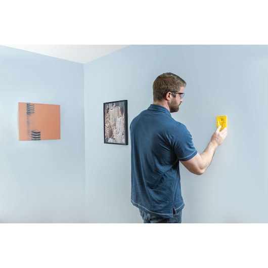 STANLEY STHT77587 stud finder held against a light blue wall, man wearing a navy shirt, indoor setting.