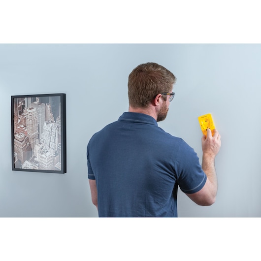STANLEY STHT77587 stud finder held against a light blue wall by a person in a navy shirt, with wall art visible.