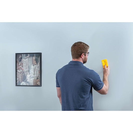 Photo of person using STANLEY STHT77587 stud finder on light blue wall near a framed cityscape picture.